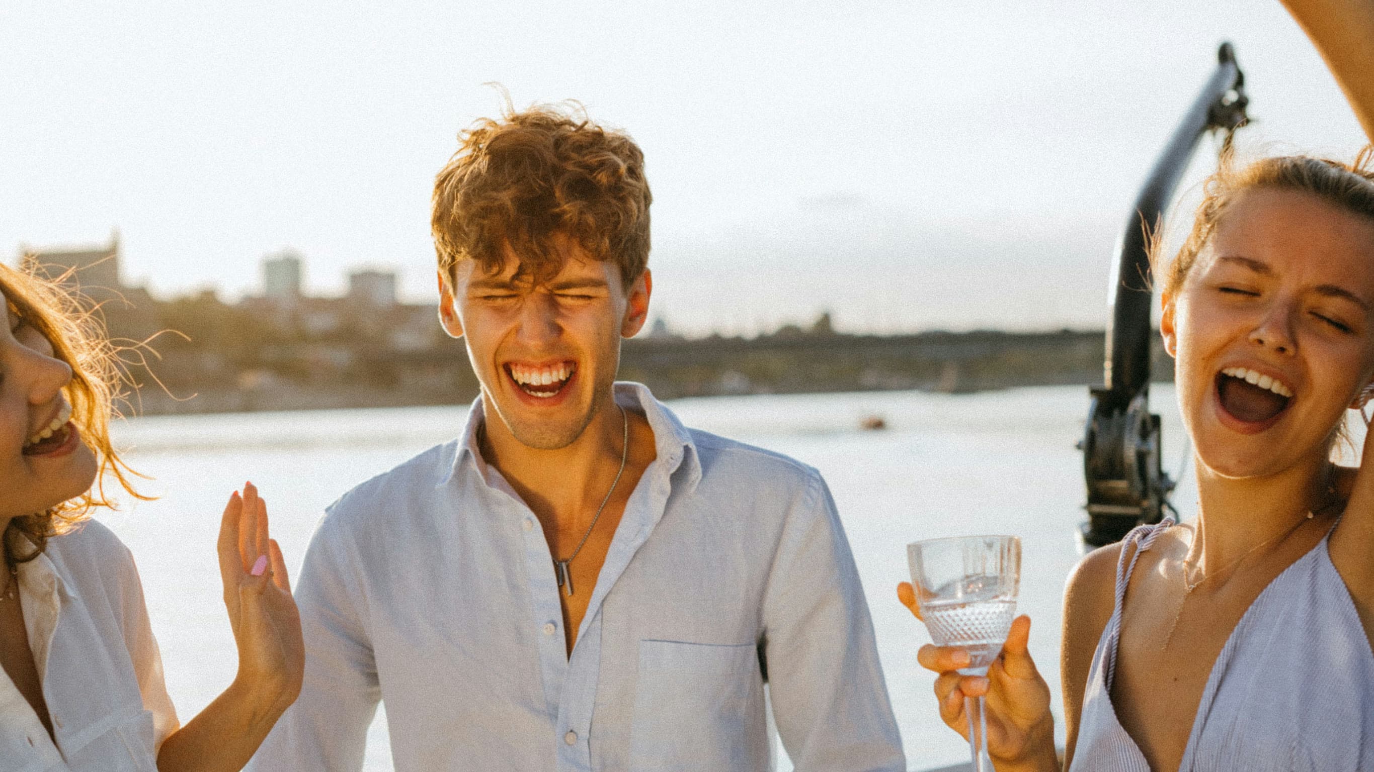 Three friends laughing on the deck of a boat at golden hour, one raising a glass, the water behind them