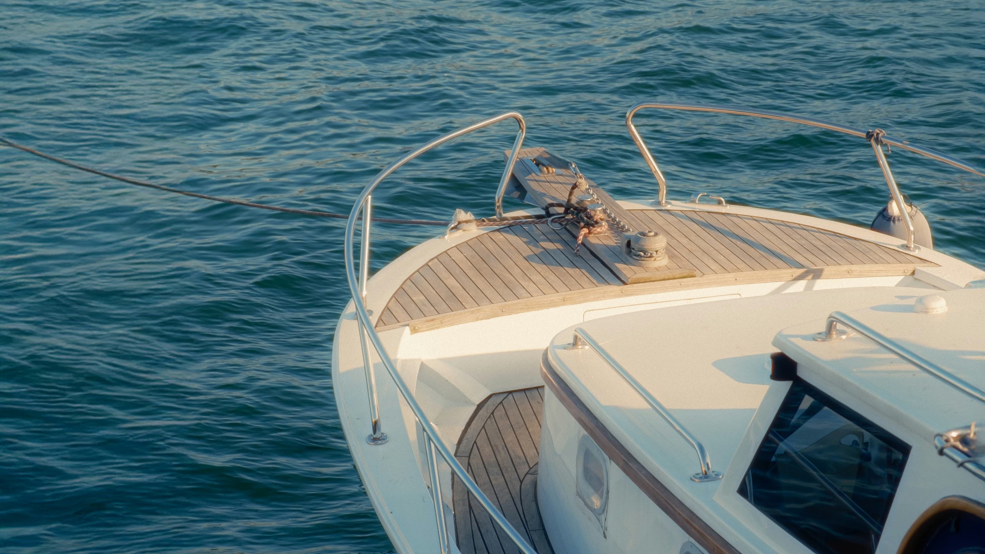 The teak foredeck and rails of a private boat at golden hour, its line running out across the water